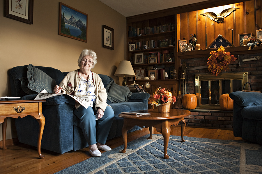 An elderly woman sits on her couch in an outdated living room smilingly lovingly