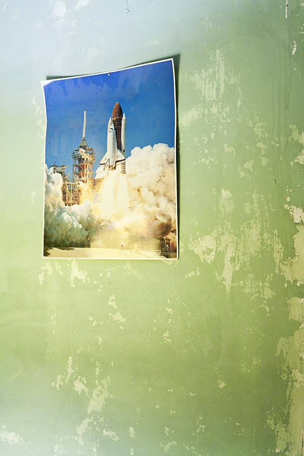 A poster of one of the NASA space shuttles blasting off sits on a light green wall with fading and peeling paint