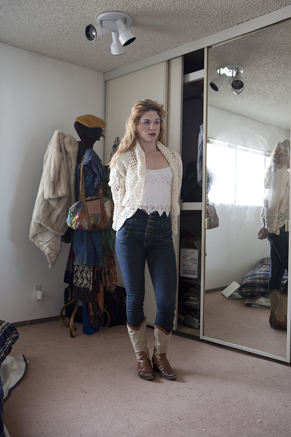 A woman stands in her bedroom wearing a faroff gaze as a part of her reflection shows in a dirty mirror