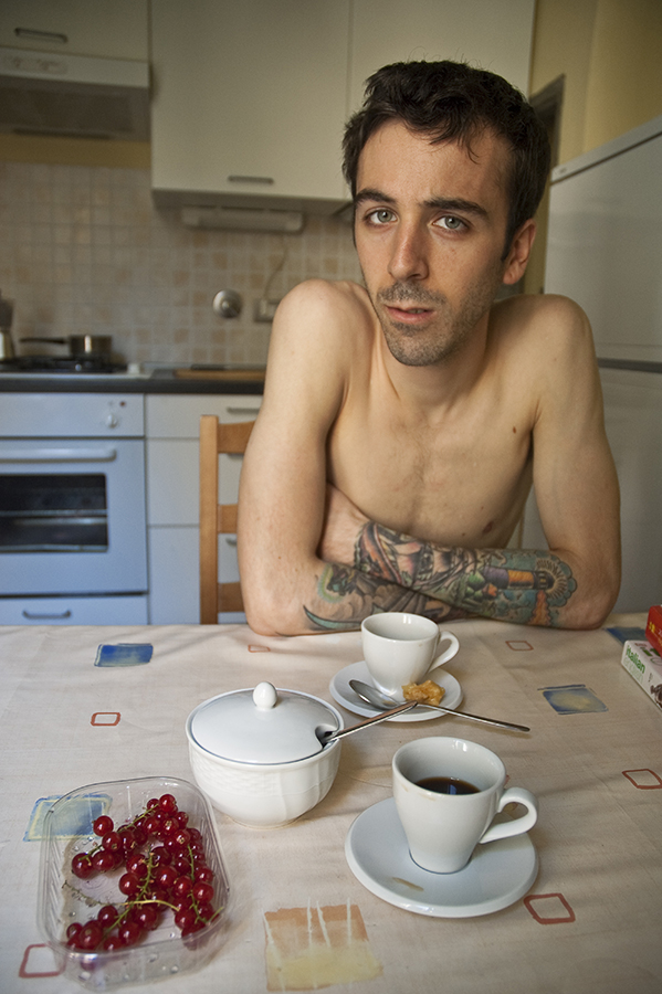 A tattooed man sits shirtless sipping an espresso at his kitchen table at his apartment in Florence, Italy