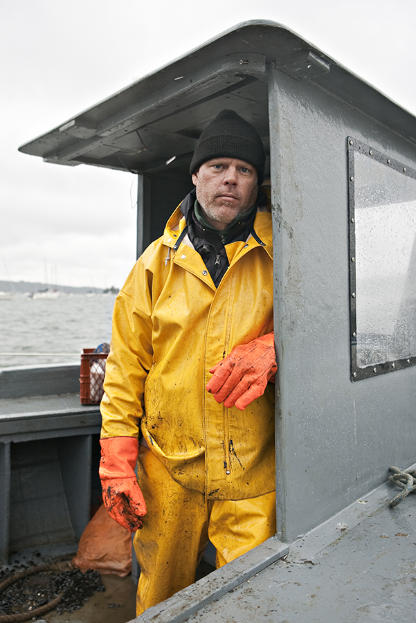 A man stands in the wheelhouse of a boat in the Long Island Sound wearing full waterproof gear including bright yellow and orange gloves